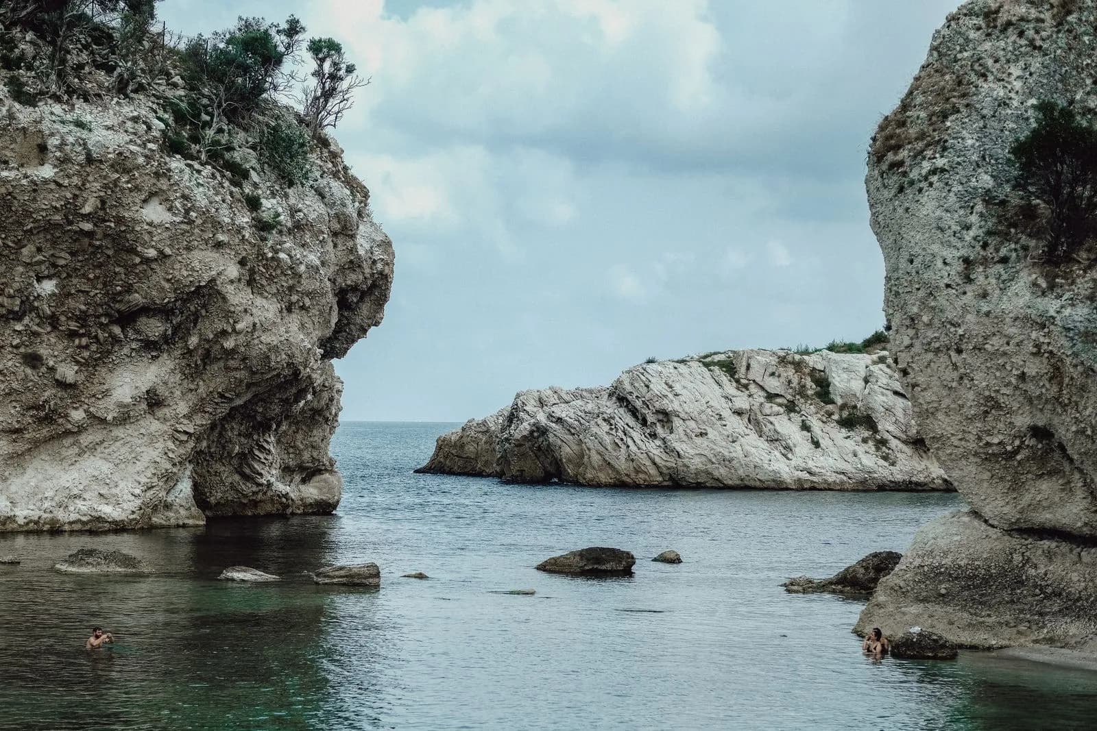 Costa rocciosa mediterranea con acqua limpida, insenatura riparata tra pareti di roccia chiara e persone in acqua