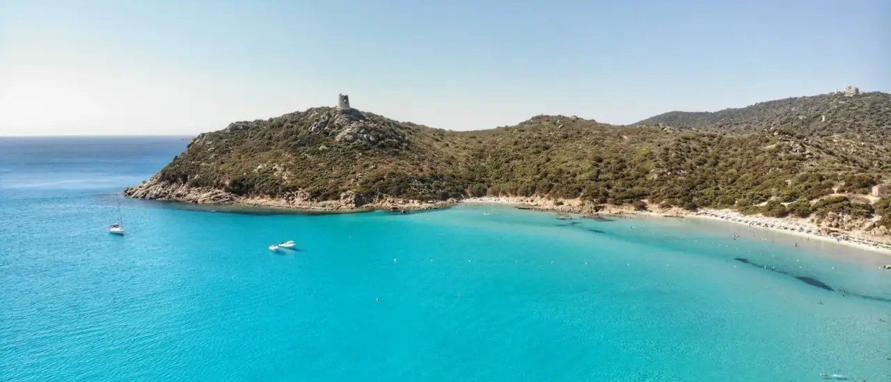 Veduta aerea della spiaggia di Porto Giunco a Villasimius, con la torre spagnola sul promontorio e barche all'ancora nell'acqua turchese